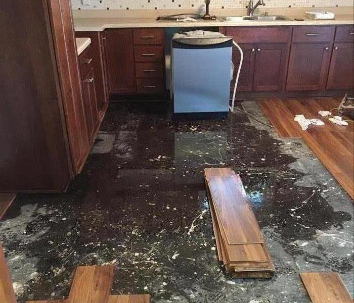 A kitchen floor covered with water and dry wood pieces removed. Also a dishwasher pulled away from kitchen counter