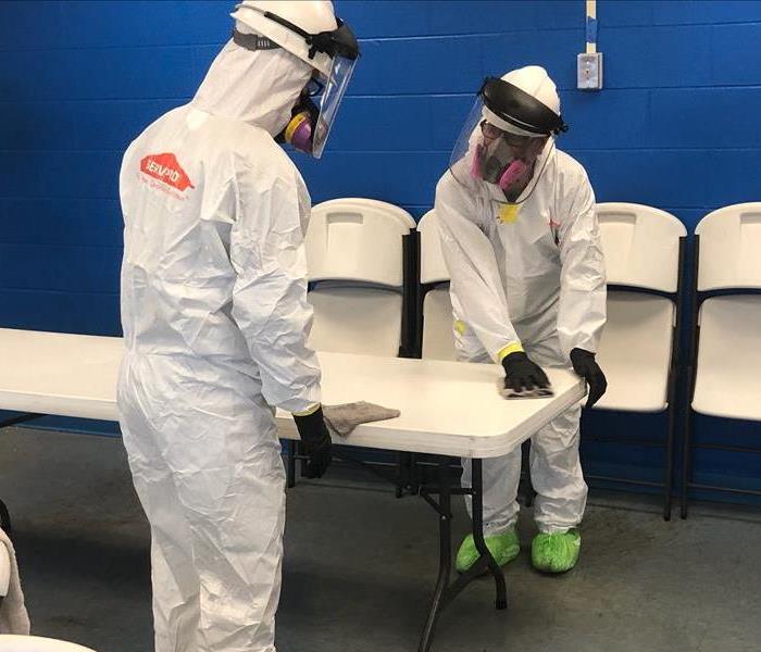 2 technicians in white personal protective equipment including facemasks and booties over their shoes cleaning a white table